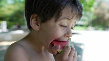 Close-up of young boy holding and eating a slice of watermelon, shirtless and focused, enjoying a refreshing summer snack, outdoor fun in the park, healthy and hydrating, playful day