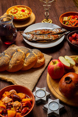 Challah, honey, apple and pomegranate and wine and chelnt and fish on the table with menorah and burning candles in honor of Rosh Hashanah