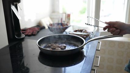 Cooking meat in a frying pan on modern stovetop, hand holding tongs ready to flip