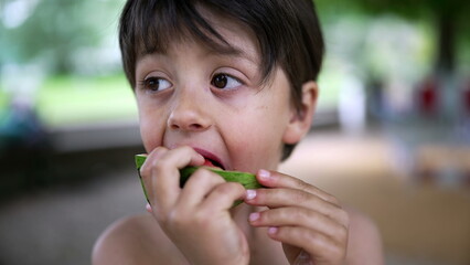 Fototapeta premium Boy savoring watermelon slice on a warm day, park setting, casual summer moment, natural backdrop, childhood joy, fresh fruit snack, healthy eating, happy and relaxed, playful expression