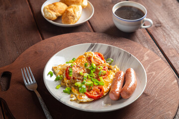 Eggs with tomatoes and fried sausage in a plate on a wooden table of coffee and croissants on a round stand
