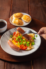Eggs with tomatoes and sausage in a plate on a wooden table of coffee and a sausage of sausage on a fork in the air above the plate