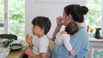 Mother drinking water while holding a baby and watching another child eat. This scene captures the...
