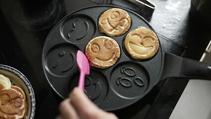 Person cooking smiley face pancakes on a stove, using a pink spatula. the fun and creativity involved in preparing a playful breakfast for a delightful start to the day