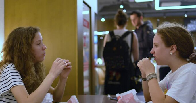 Curly haired teenage girls eat sandwiches sitting together in airport cafeteria before flight. Pretty sisters have lunch in restaurant for passengers