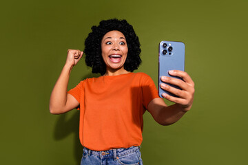 Photo of pretty young woman smart phone selfie raise fist wear orange t-shirt isolated on khaki color background