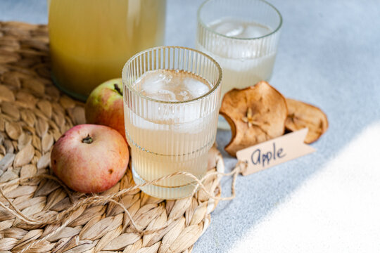 Jug and two glasses of fresh apple juice on a table mat with fresh apples, dried apple slices and a tag with the word apple