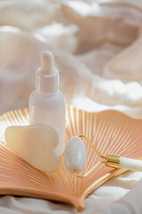 White jade face roller and gua sha massager, bottle of serum or essential oil on sunlit background with shadow.