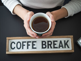A woman holding a cup of hot coffee in her hand, with "Coffee Break" written on the work desk table, the photo is taken from above