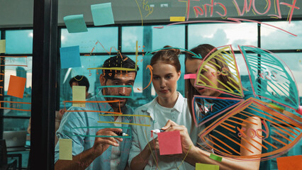 Professional business team brainstorm, sharing, thinking idea while skilled male leader write on sticky notes at glass wall. Skilled manager placed business plan on glass wall at office. Tracery