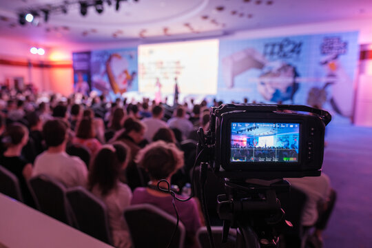 Professional video camera in foreground recording conference with engaged audience and colorful stage lighting
