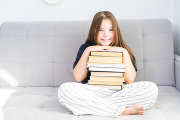 Little pre-teen girl holding a stack of books, putting her hands and head on it, sitting on a gray...