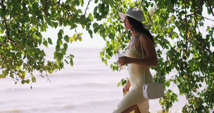 Elegant woman in dress and hat with cocktail walking at the river beach