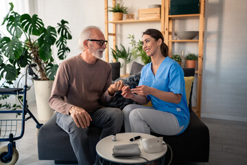 A smiling female nurse in blue scrubs is attentively measuring the blood sugar levels of a content elderly gentleman in his cozy apartment, with medical tools nearby.