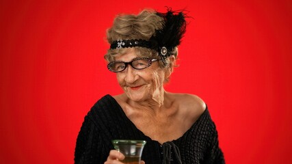 Smiling portrait of fun elderly old woman drinking wine and smiling wearing wig and glasses isolated on red background.