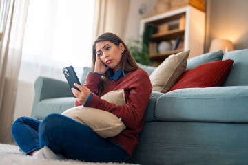 Obraz premium An uneasy Caucasian woman lounges in a living room, tightly hugging a pillow as she peers at her smartphone, dressed in casual home attire.