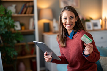 A cheerful woman holds a credit card and digital tablet, ready for an online purchase from her...