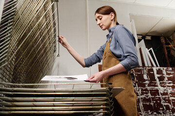 Low angle of young craftswoman with sketch in hand standing by printing equipment with flap lid and...