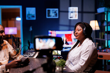 During a vlog recording session, a man is using a professional microphone and audio mixer while interviewing an African American female influencer in front of a digital camera.