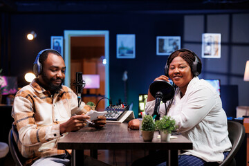 Black journalist uses modern recording technology to interview smiling female content creator in home studio. They broadcast their lively conversation to online audience via social media platforms.