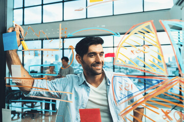 Smiling smart businessman writing while thinking about business plan while standing at financial statistic chart on glass wall.Professional manager planing, designing about business idea. Tracery.