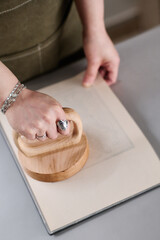High angle of hands of young female designer pressing round wooden tool to paper covering workpiece with artwork pattern