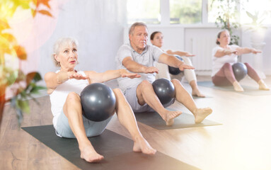 Fototapeta premium In fitness studio, people of different ages engaged in Pilates. Young girl coach performs exercise with softball with elderly people. They sitting on floor with legs bent and holding softball in knees