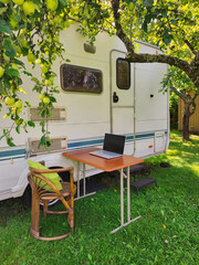 Serene outdoor office setup with a laptop on a wooden table beside an RV and under apple trees.