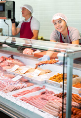 A smiling, friendly young saleswoman working behind the counter of a butcher shop takes out delicious fresh baby chorizo sausages from a glass case and demonstrates them..