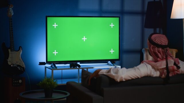 Arab man lounged on cozy sofa, watching news broadcast on green screen device while relaxing at home. Muslim person chilling on couch, looking at mockup TV channel showing current events