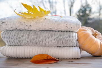 stack of soft, knitted sweaters in autumnal colors displayed alongside pumpkins and leaves,...