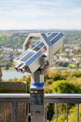 A telescope facing the scenic view of Schloss Drachenburg, offering a breathtaking panorama of the surrounding landscape with greenery and distant river.