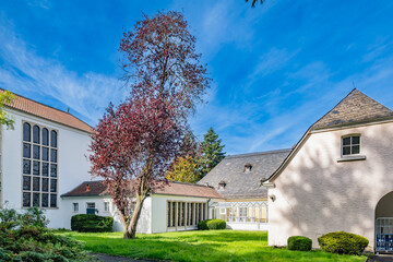A serene courtyard area of Klosterruine Heisterbach with manicured green lawns, surrounding trees, and buildings with white facades under a bright blue sky.