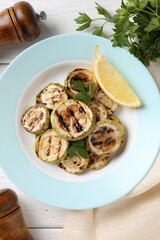 Tasty grilled zucchini slices with lemon and parsley on white wooden table, flat lay