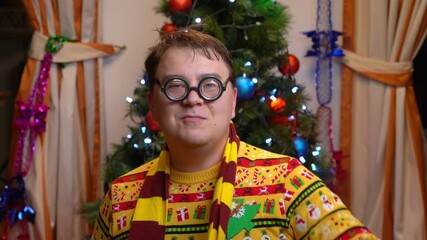 Portrait of funny man in bright festive clothes, ugly sweater, round nerd glasses, congratulatory speech, defiantly wrapping striped scarf around neck Comical address from blogger at Christmas tree - Powered by Adobe