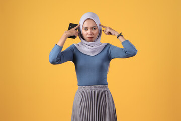 pensive asian indonesian muslim women holding smart phone with thinking gesture on isolated yellow background