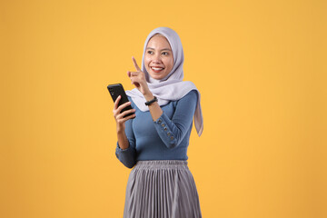 happy asian indonesian muslim woman holding smart phone with pointing finger gesture on isolated yellow background