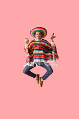 Obraz premium Handsome young man in colorful poncho, sombrero and with sugar skull jumping on pink background