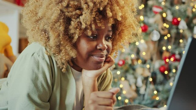 Happy African American woman lying on bed, smiling and typing message on laptop in cozy room, Christmas tree decorated with fairy lights in the background