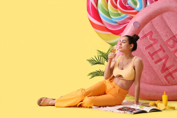 Young woman with cocktail, palm and inflatable mattresses sitting on yellow background