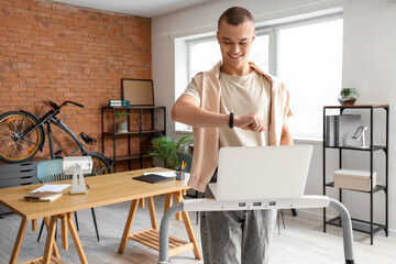 Young businessman with laptop looking at smartwatch on treadmill in office