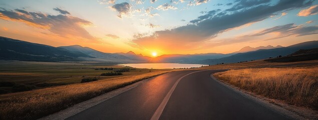 Panoramic view of a winding road leading to the horizon with mountains, a lake, and a vibrant sunset sky with clouds, offering a serene and picturesque landscape.