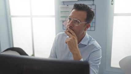 Hispanic man thinking and discussing ideas in an office workspace with large windows and motivational notes on the wall.
