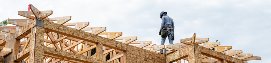 New residential home construction in framing stage, workman with hardhat and toolbelt on the roof rafters studying next piece of work, OSB, oriented strand board, sheathing on exterior walls with wind © knelson20