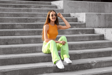 Beautiful young woman in stylish sunglasses sitting on stairs outdoors