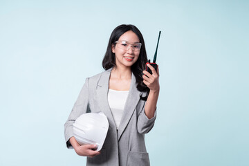 Confident woman engineer holding walkie talkie and white helmet isolated on light blue background.