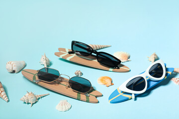 Different stylish sunglasses with surfboards and seashells on blue background