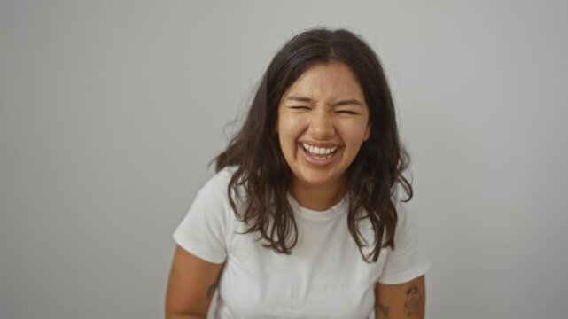 Woman standing and laughing hard, young hispanic woman wearing white t-shirt, smiling loudly over isolated background due to funny joke, enjoying fun and out loud laughter