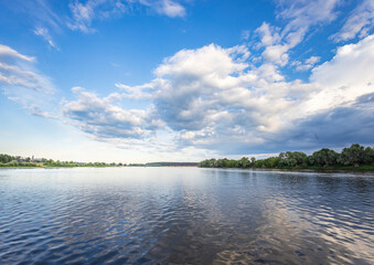 A wide, calm river stretches out, reflecting the blue sky and puffy white clouds above. Lush green...