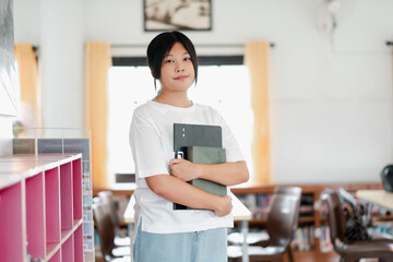 A young student stands in a bright classroom holding notebooks, with bookshelves and chairs visible in the background.